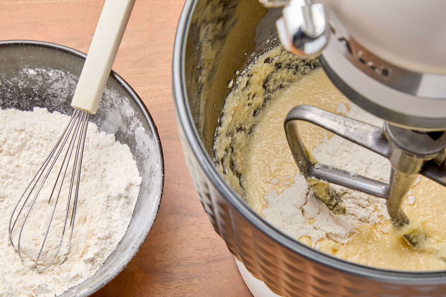 Three-quarter shot of a bowl of flour beside a stand mixer, which is on and mixing the batter with the paddle attachment. A couple of lumps of flour remain unincorporated in the batter. The mixer is positioned on a wooden countertop.