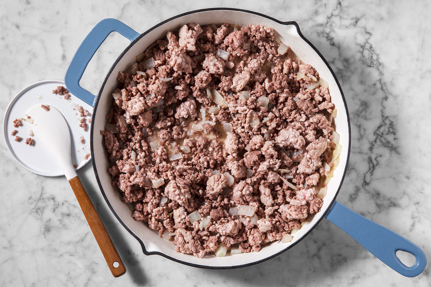 A pan with browned ground beef on a marble countertop. A white spatula with a wooden handle rests nearby.