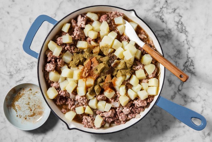 A blue-handled pan on a marble surface contains diced potatoes and ground beef with a sprinkle of spices. A wooden-handled spatula rests on the pan's edge. A small bowl with spices is next to the pan.