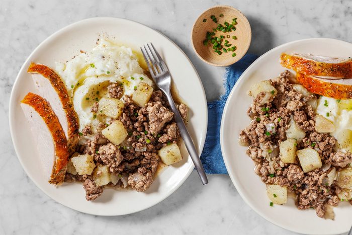 Two plates of a savory meal, each with seasoned ground meat, diced potatoes, and fluffy mashed potatoes topped with chives. A few strips of roasted vegetables are on the side. A small bowl of chives nearby. Forks rest on the plates.