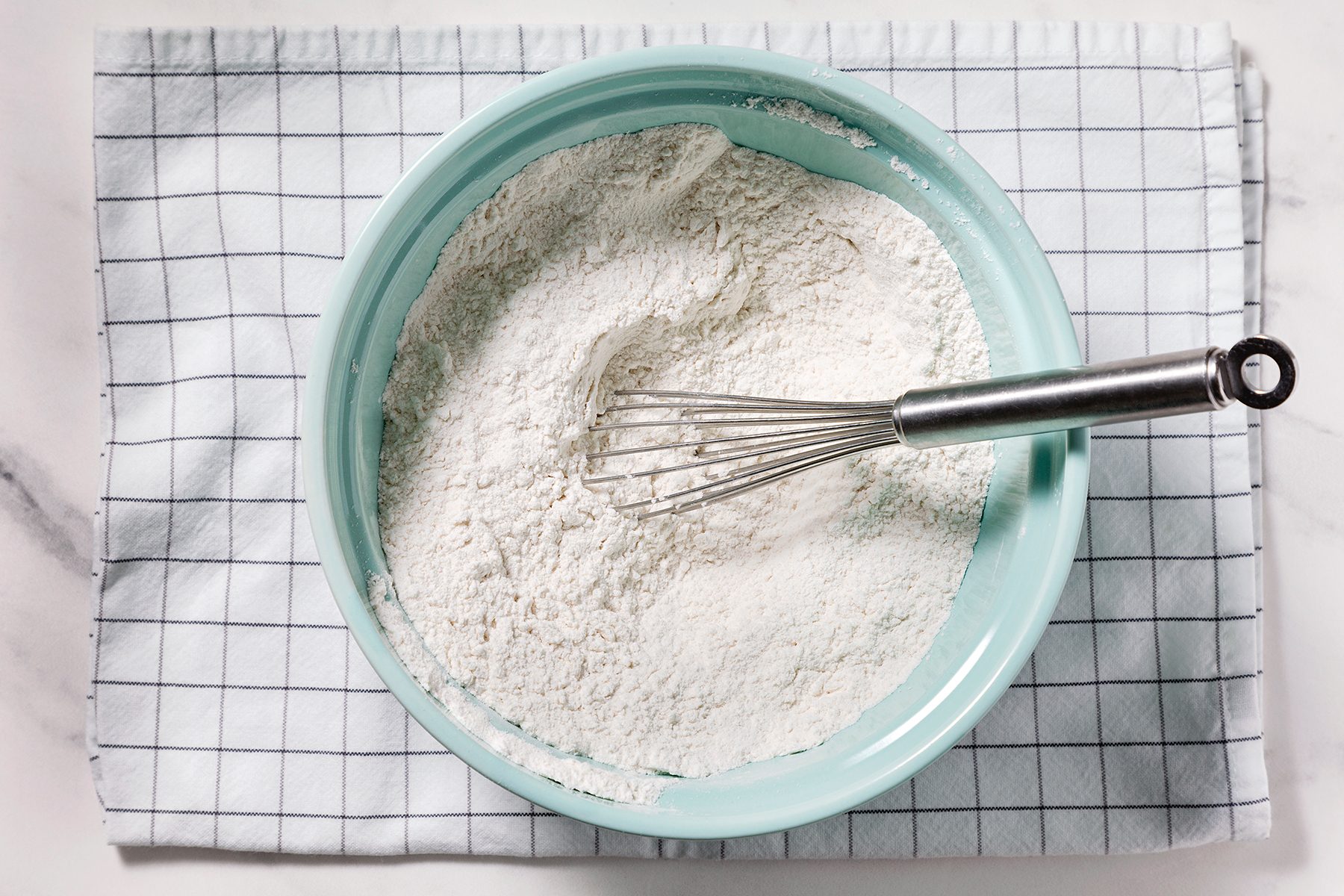 A light blue bowl filled with flour and a metal whisk rests on a white and blue checkered cloth, placed on a marble countertop.