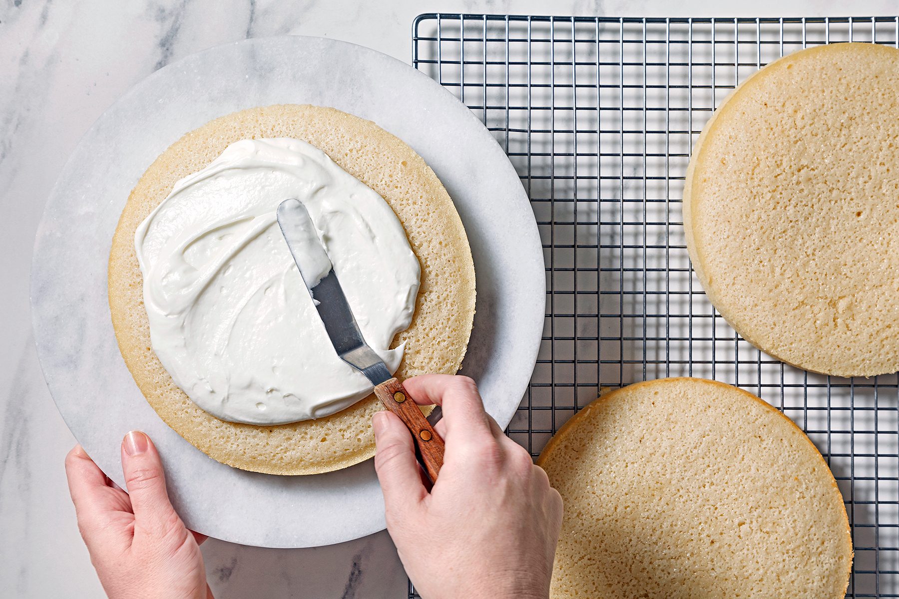 A person spreads white frosting on a round cake layer using a spatula. The cake is placed on a marble surface, and two more cake layers are nearby on a cooling rack.