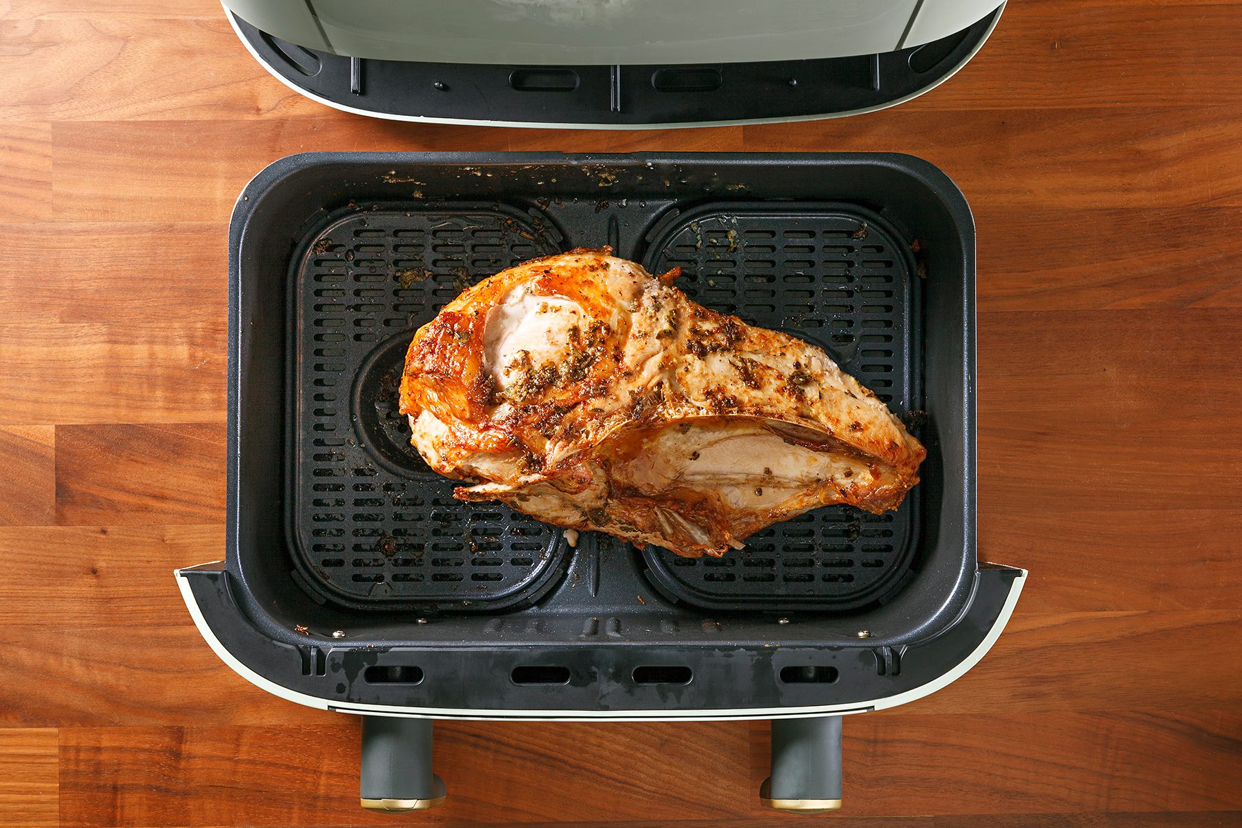 A cooked piece of meat is inside an open air fryer on a wooden surface. The meat is seasoned and browned, resting on the fryer basket. The top of the air fryer is visible.