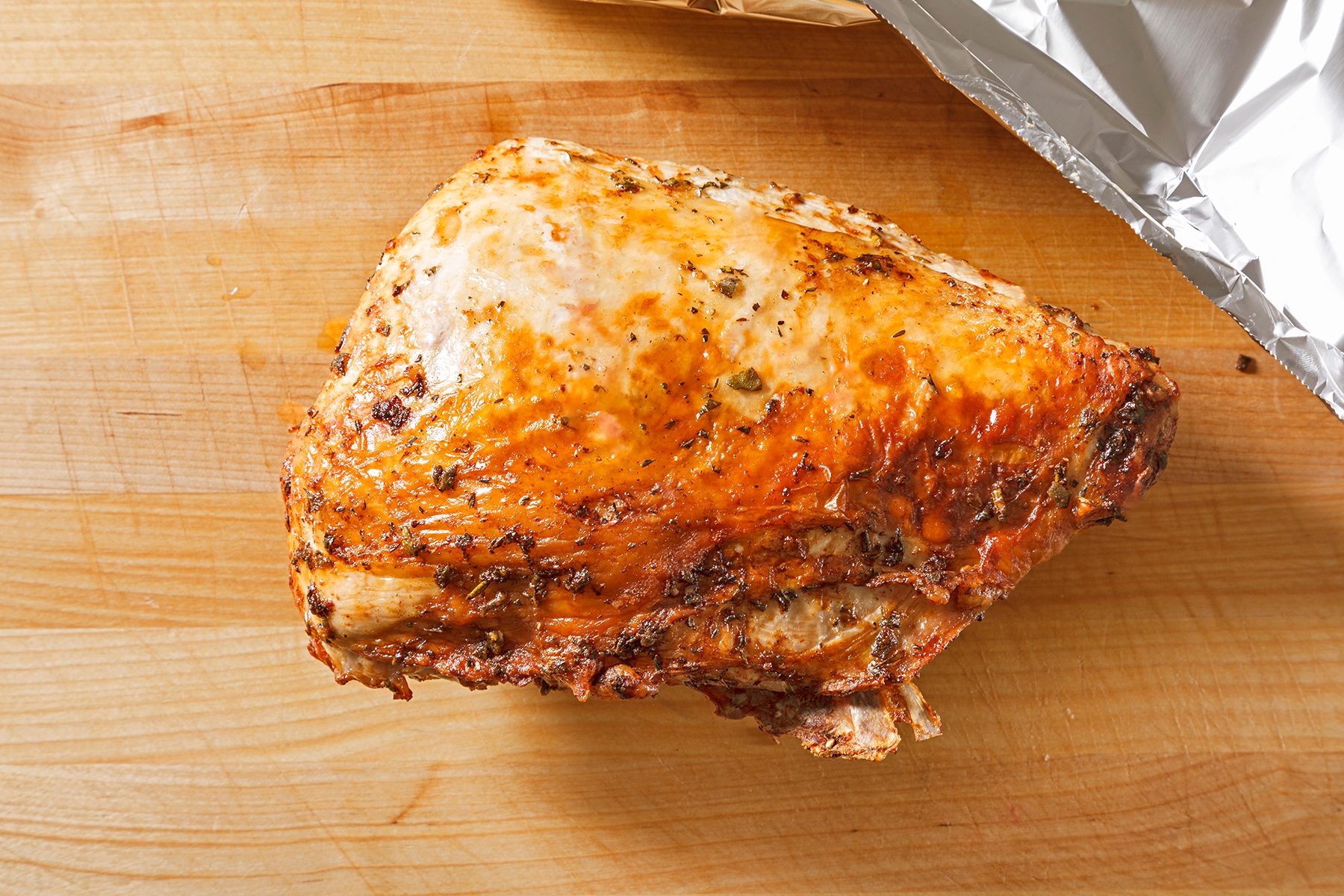 A seasoned and cooked chicken breast with a crispy skin rests on a wooden cutting board. A piece of aluminum foil is partially visible in the top right corner.