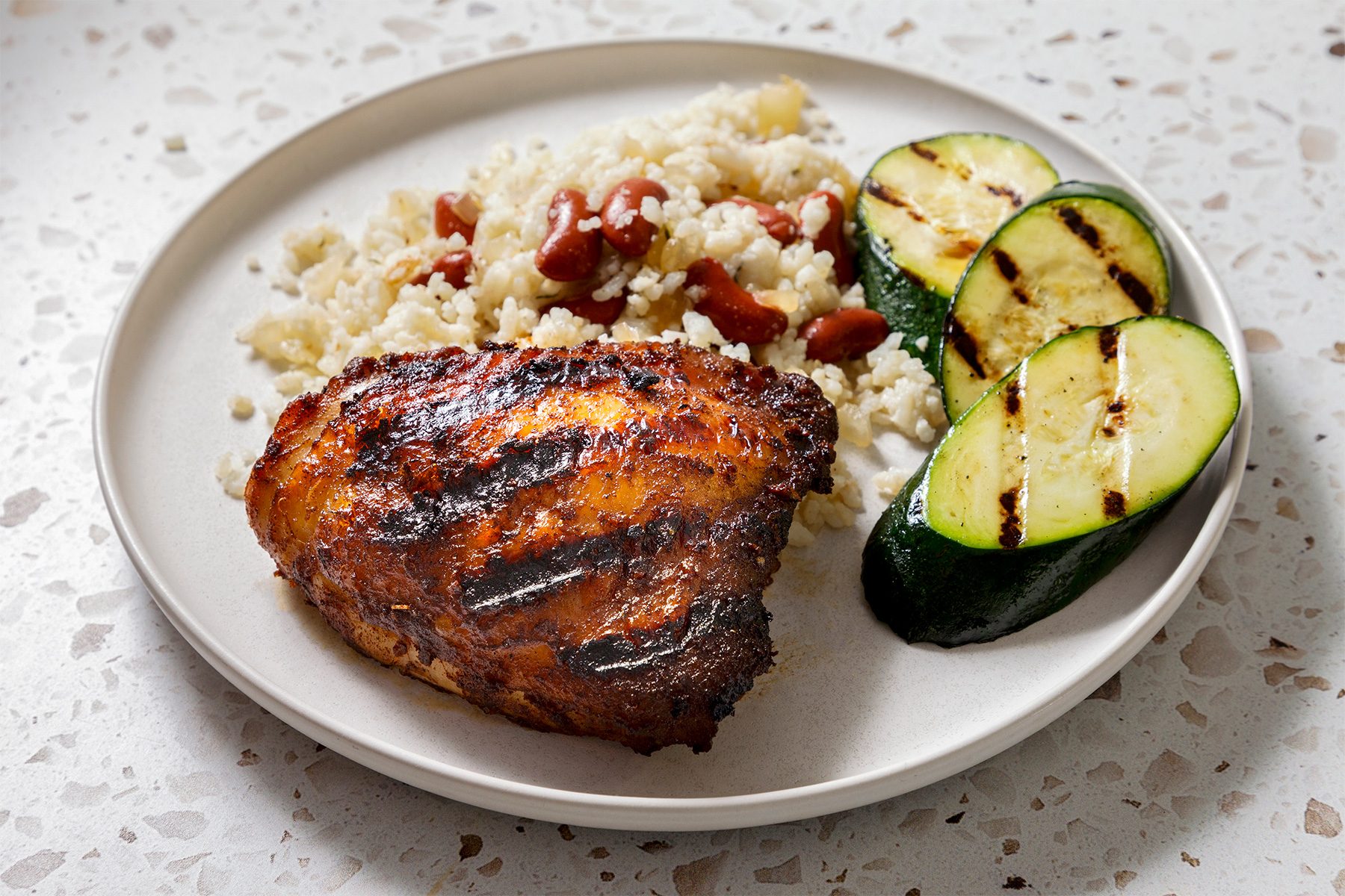 3/4th shot of a plate of food with grilled chicken, rice, red beans and zucchini, and a plate of grilled chicken with a small bowl of red sauce, a fork, and a few drops of sauce on the plate, all laid out on a white countertop with brown flecks