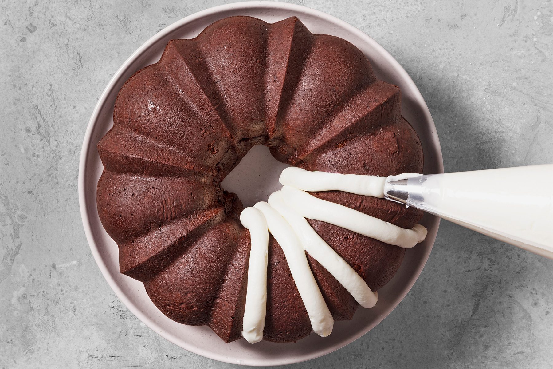overhead shot of a chocolate bundt cake with white frosting being piped onto it, The cake is sitting on a pink plate and the plate is sitting on a gray textured surface, The frosting is being piped in a spiral pattern around the cake, The cake is almost entirely covered in frosting, The piping bag is visible in the upper right corner of the image