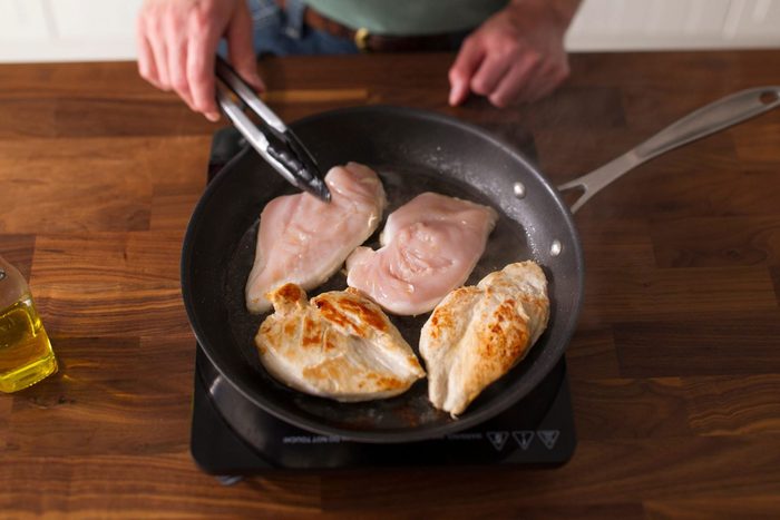 A person is using tongs to cook chicken breasts in a frying pan on an induction stovetop. Two pieces are raw while two are golden brown. A bottle of olive oil sits nearby on a wooden countertop.