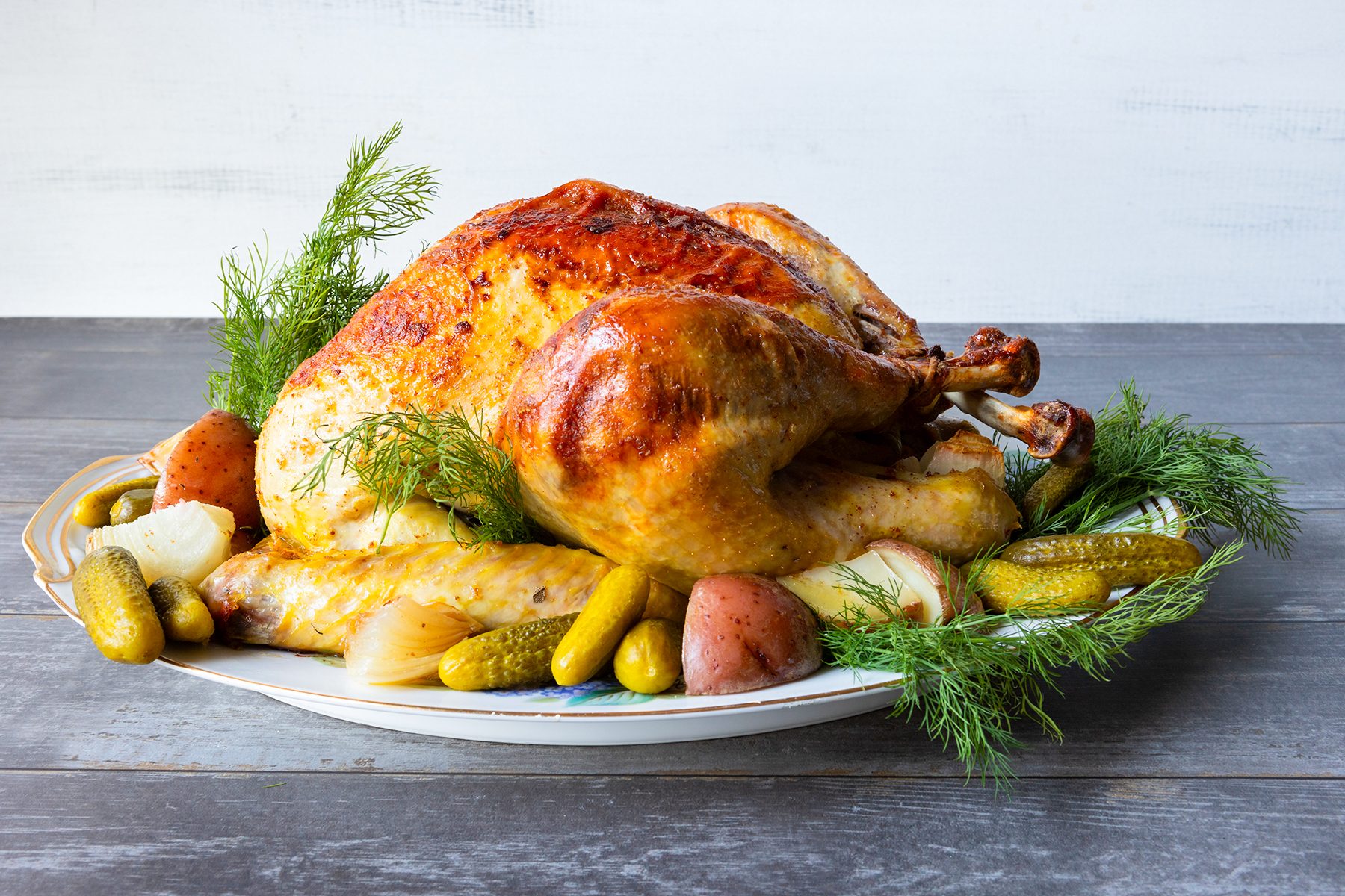 A roasted turkey on a platter, surrounded by dill, small potatoes, and pickles. The turkey has a golden brown, crispy skin, and the platter is placed on a wooden surface.