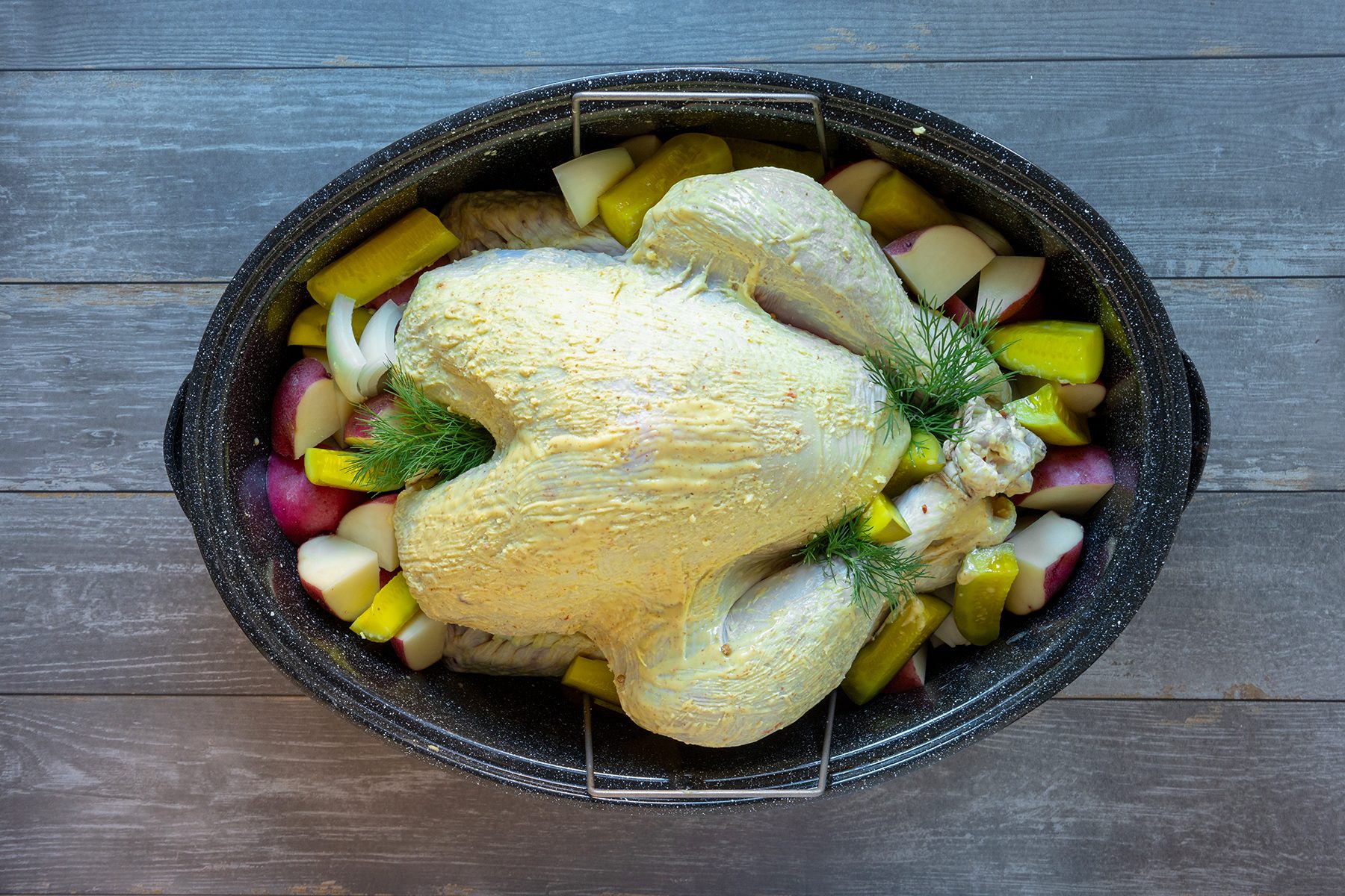 A seasoned raw turkey is placed in a roasting pan, surrounded by assorted chopped vegetables like potatoes and zucchini, garnished with fresh herbs, ready for cooking on a wooden surface.
