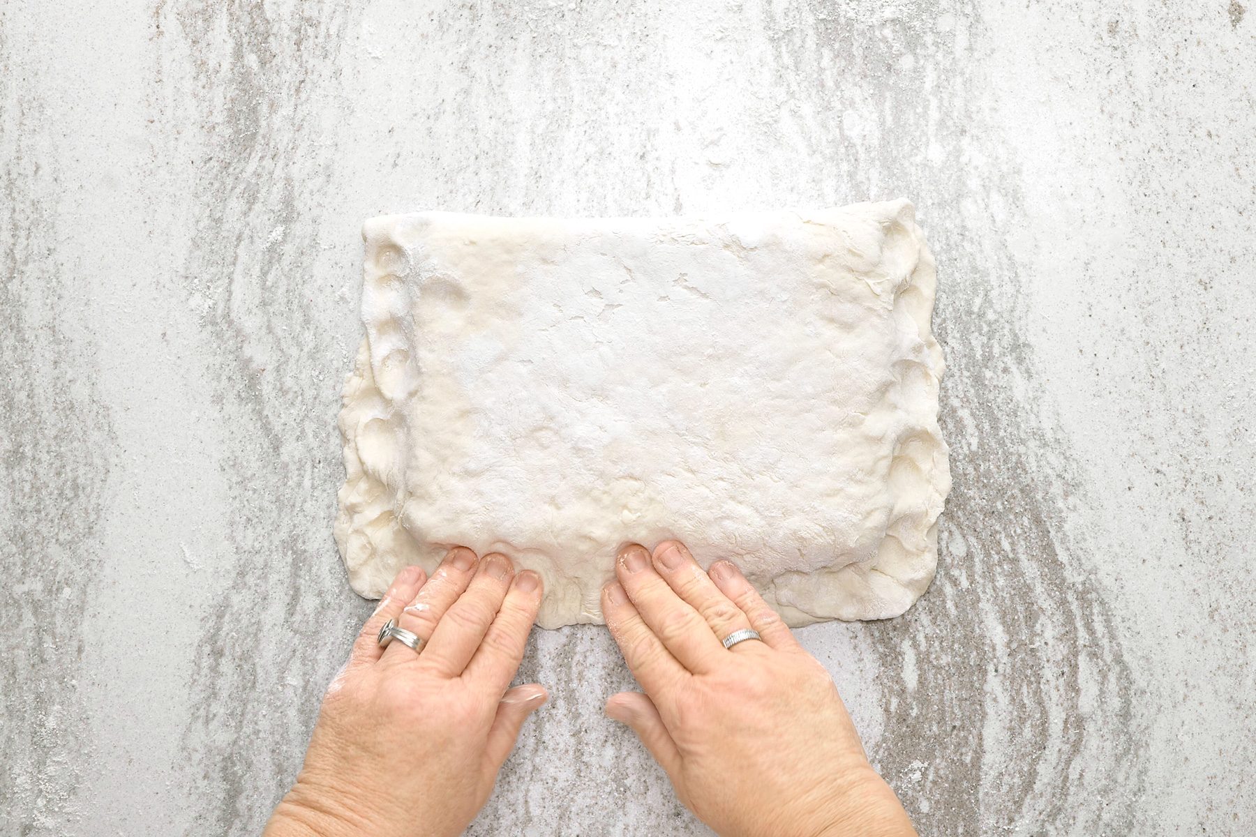 overheda shot of a person's hands, pressing down on a square of dough, possibly pie crust, with both hands; the dough is dusted with flour, it is sitting on a light gray counter or table, there is flour scattered around the dough;