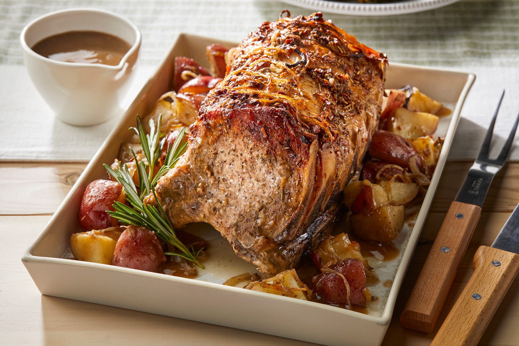 Horizontal AP; Pull shot of a roasted pork with potatoes and onions in a white baking dish; the dish is sitting on a white linen napkin and a light brown wood table; there is a sprig of rosemary on top of the pork; there is also a white bowl of gravy next to the baking dish; there are two wooden handled knives and a fork sitting to the right of the baking dish