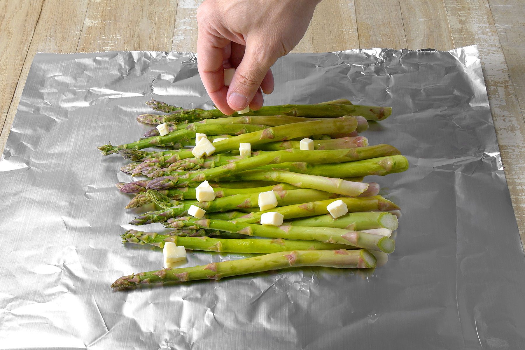 A hand is sprinkling small cubes of butter over a bunch of fresh green asparagus spears arranged on a sheet of aluminum foil. The foil is placed on a wooden surface.