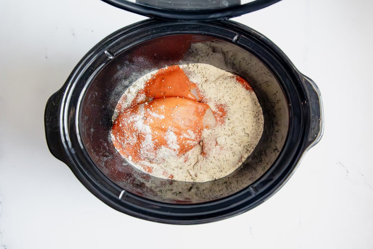 Overhead shot for Taste of Home Shredded Buffalo Chicken with chicken, buffalo sauce, and ranch seasoning in a large slow cooker.