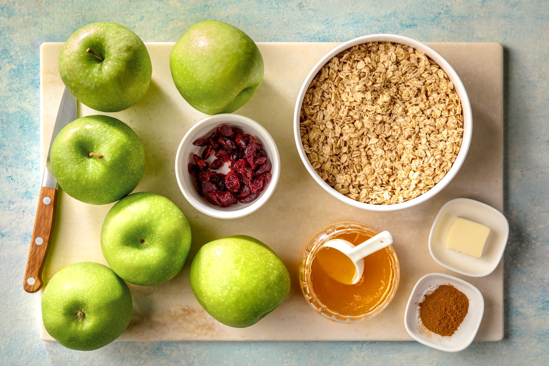 overhead shot of ingredients for a healthy baked apple oatmeal; the ingredients include six green apples, a knife, a bowl of oats, dried cranberries, a dish of honey, a small dish of butter, and a dish of cinnamon powder; The ingredients are arranged on a marble cutting board, which is placed on a light blue background