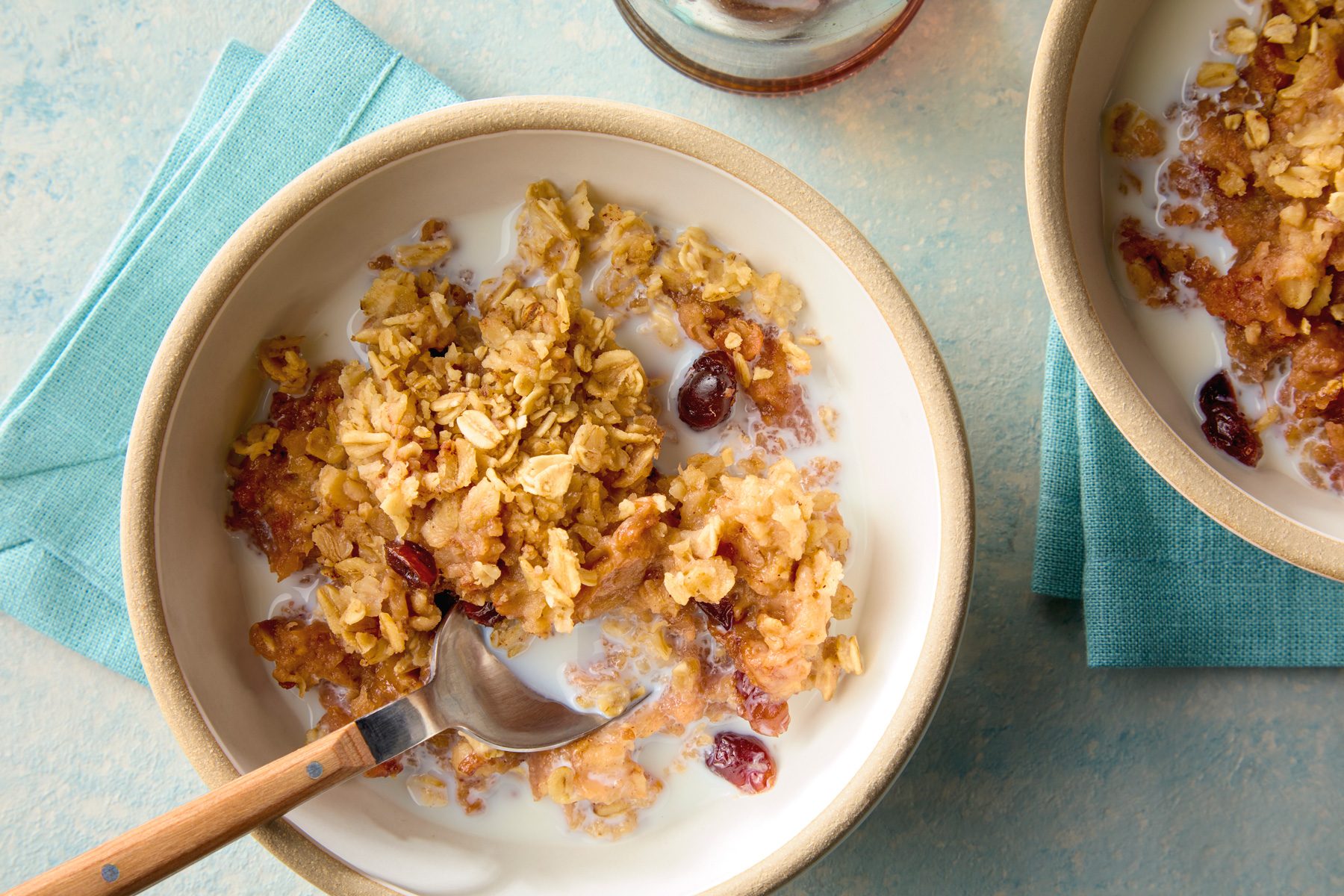 overhead shot of a bowl of oatmeal with milk and fruit; it is on a blue napkin, a wooden spoon is in the bowl; there is another bowl of oatmeal in the background on another blue napkin; the background is a blue speckled surface