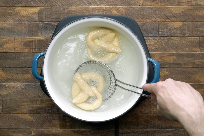 A hand holds a wire skimmer lifting a soft pretzel from a pot of boiling water. Another pretzel is in the water. The pot sits on a wooden surface.