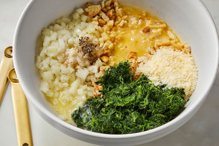A large white bowl containing chopped onions, bread crumbs, a mixture of beaten eggs and seasonings, grated cheese, and chopped fresh herbs. Two gold measuring spoons are placed beside the bowl on a light surface.