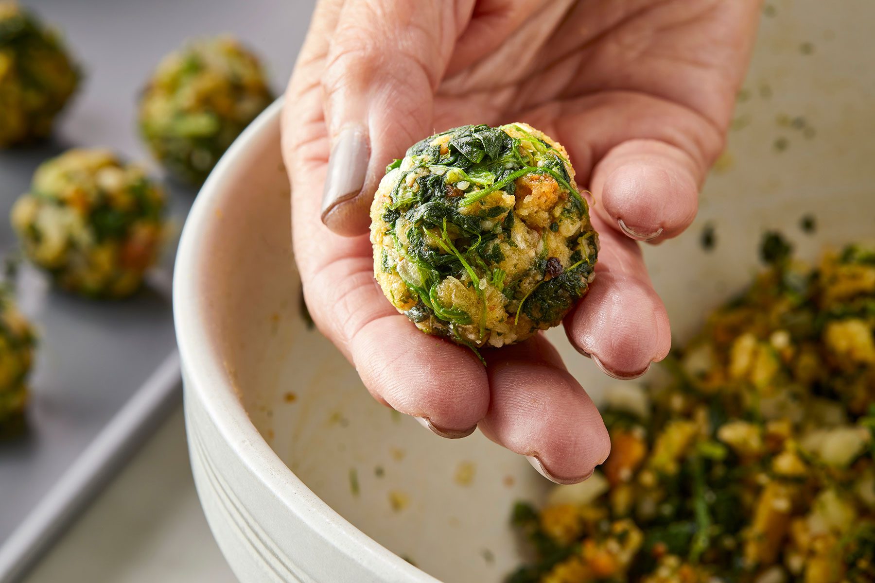 A hand holds a freshly made spinach ball over a mixing bowl. The spinach ball is packed with visible green spinach pieces and other ingredients. More spinach balls are on a tray in the background.