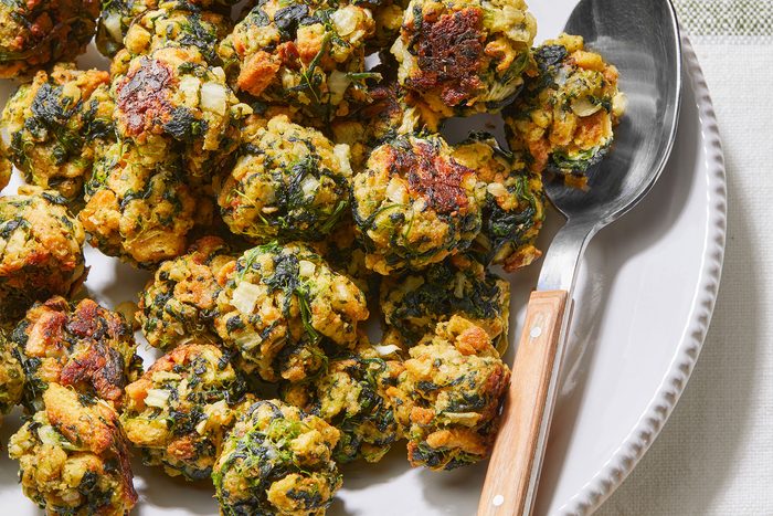 A plate of golden-brown spinach and herb fritters, slightly crispy on the outside, with a serving spoon placed on the right edge of the plate. They are arranged on a light-colored tablecloth.