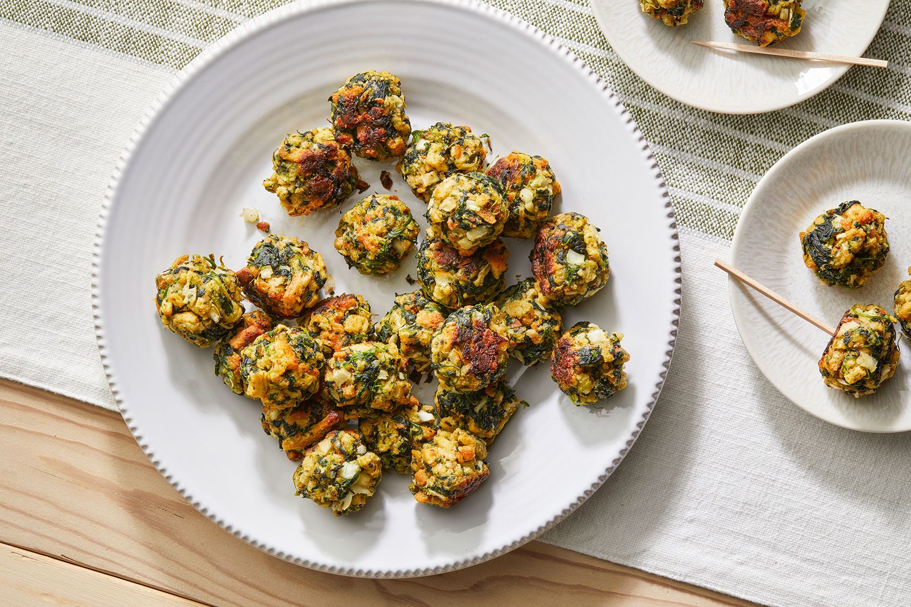 A large white plate filled with spinach and cheese balls, arranged on a light wooden table with a striped tablecloth. Small skewers are beside a smaller white plate holding a few of the balls.