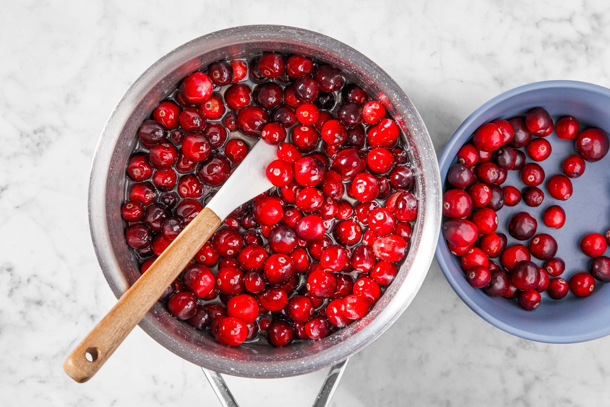 A pot filled with glistening red cranberries and a wooden spoon sits on a marble countertop. Next to it is a blue bowl containing additional cranberries.