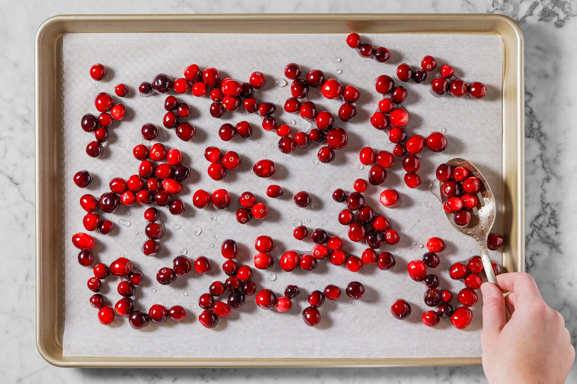 A baking tray lined with parchment paper is spread with fresh cranberries. A hand in the lower right corner uses a spoon to arrange the cranberries. The tray rests on a marble countertop.