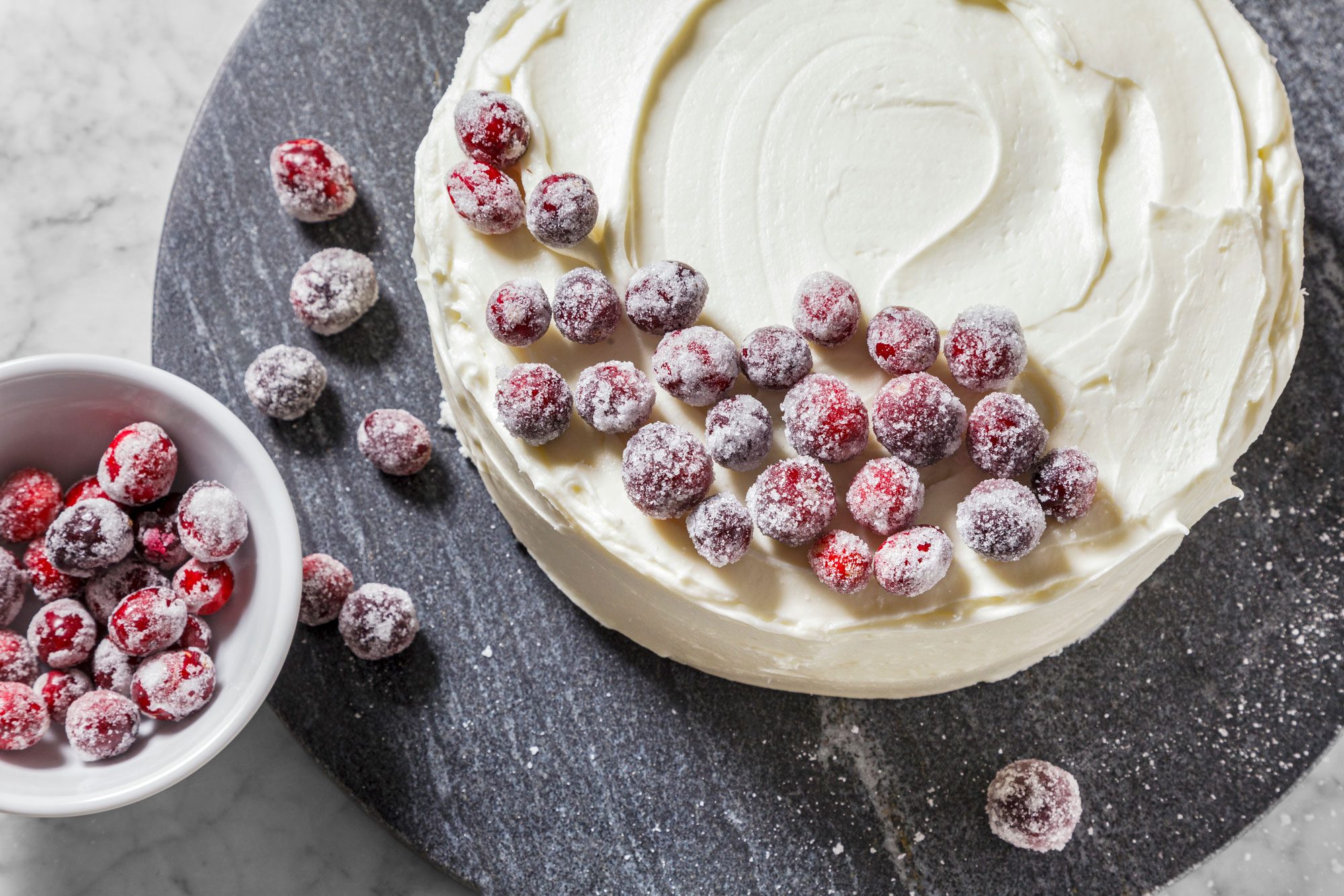 A round cake with smooth white frosting is topped with sugared cranberries. A small bowl next to the cake holds more sugared cranberries. The cake sits on a dark slate surface.