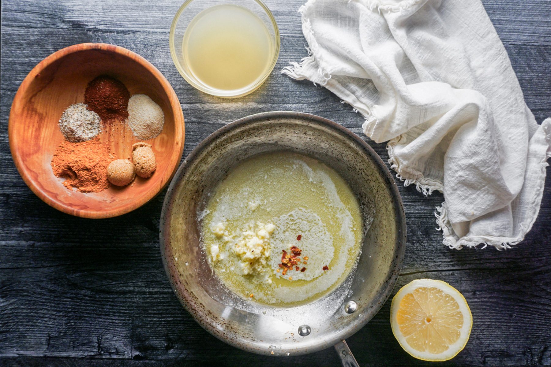A pan with a green sauce, garlic, and chili flakes on a dark table. A halved lemon, a bowl of mixed spices, a glass of juice, and a white cloth are placed nearby.
