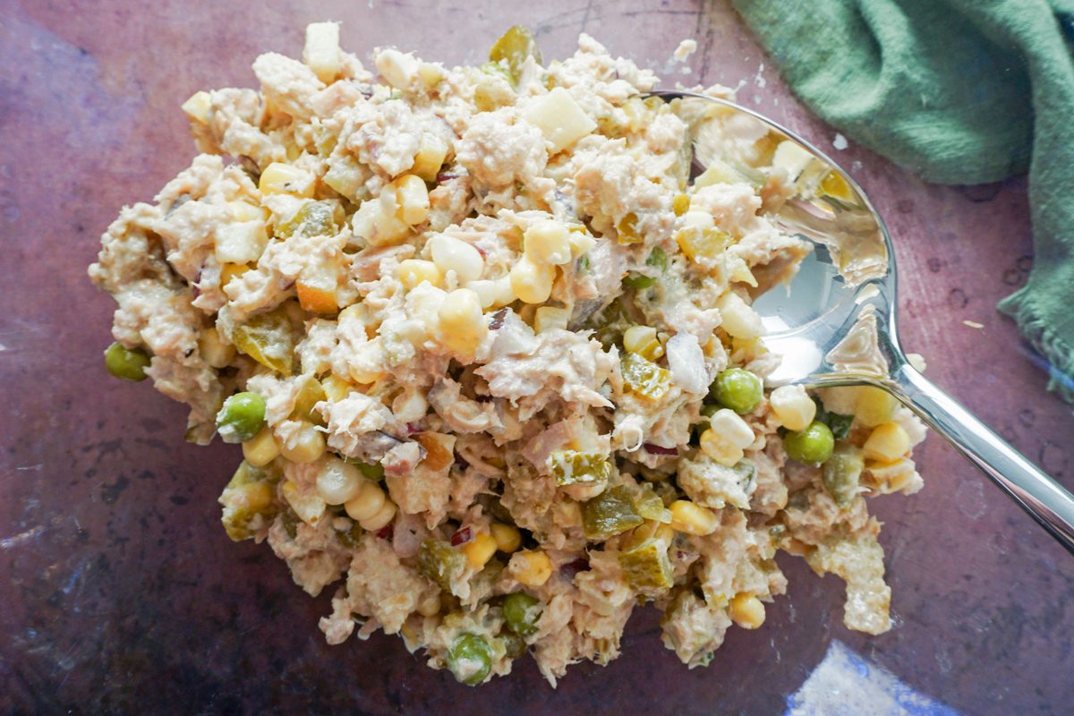 A serving of tuna salad with corn, green peas, and diced pickles on a spoon sits on a textured surface. A green cloth is partially visible in the background.