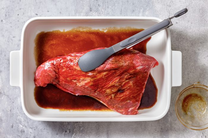 Raw beef marinating in brown sauce in a white dish, with tongs resting on top