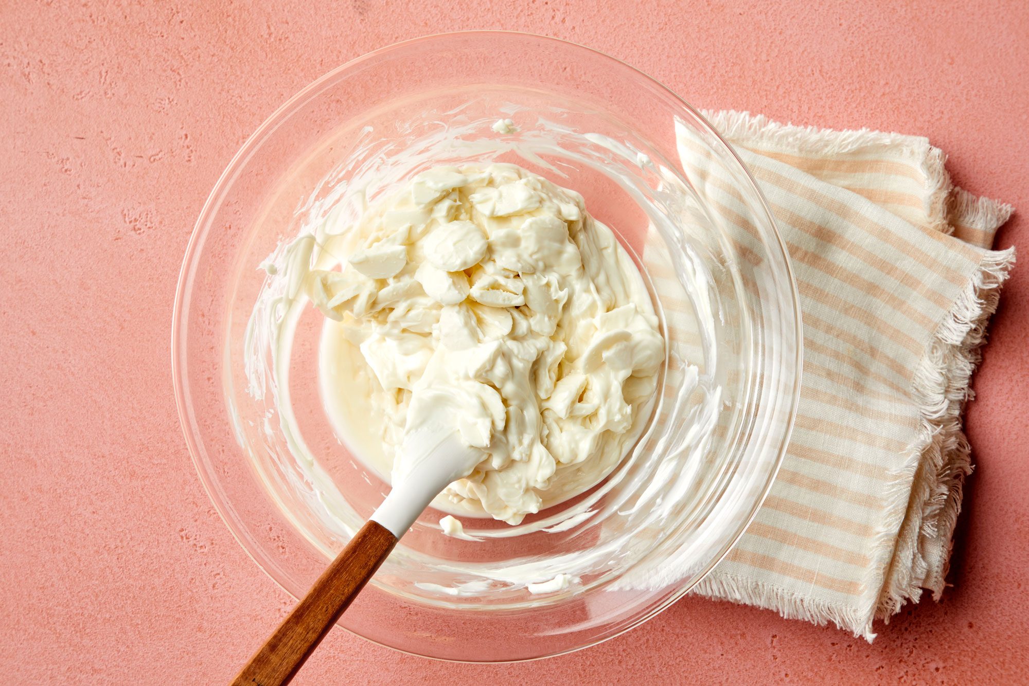 overhead shot of melted candy coating; stir until smooth in a glass bowl placed over pink background, a kitchen towel is placed on the right side of the bowl;