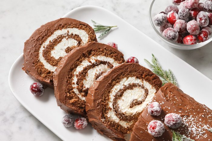 Close shot of Chocolate Yule Log; serve on white tray; dust with additional confectioners sugar and decorate with sugared cranberries and sugared rosemary sprigs; cut into pieces; marble surface.