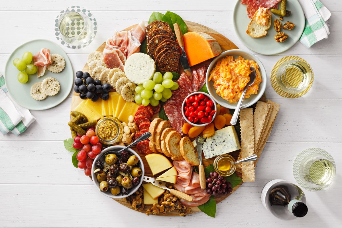 A round charcuterie board with assorted cheeses, meats, olives, nuts, crackers, and fruits. Three plates and glasses of white wine are arranged on a white wooden table.