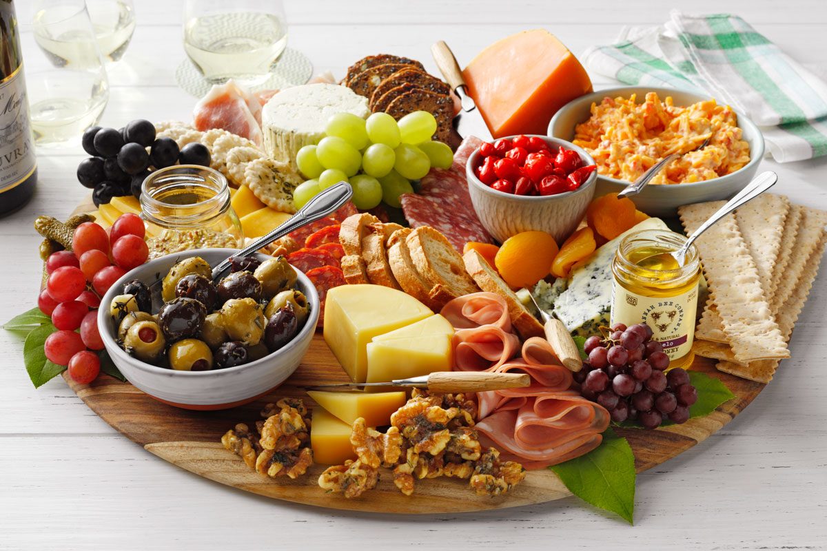 A colorful charcuterie board featuring assorted cheeses, meats, grapes, olives, cherry tomatoes, crackers, nuts, and pomegranate seeds. A glass of white wine and a checkered napkin are in the background.