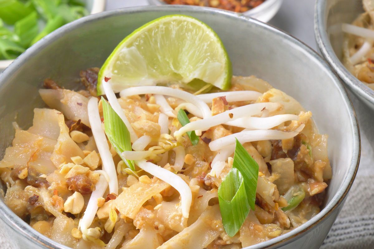 a close-up view of a bowl of Chicken Pad Thai; the noodles are topped with bean sprouts, crushed peanuts, and chopped scallions; a wedge of lime sits on top of the dish; the bowl is sitting on a white linen napkin