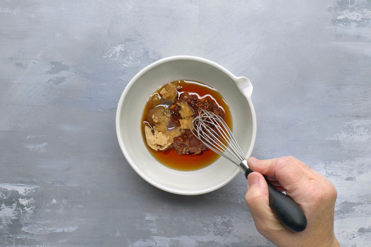 overhead shot of a hand holding a whisk in a white bowl filled with a sauce mixture; The sauce is a light brown color and contains brown sugar and chili flakes; The bowl is sitting on a grey textured surface