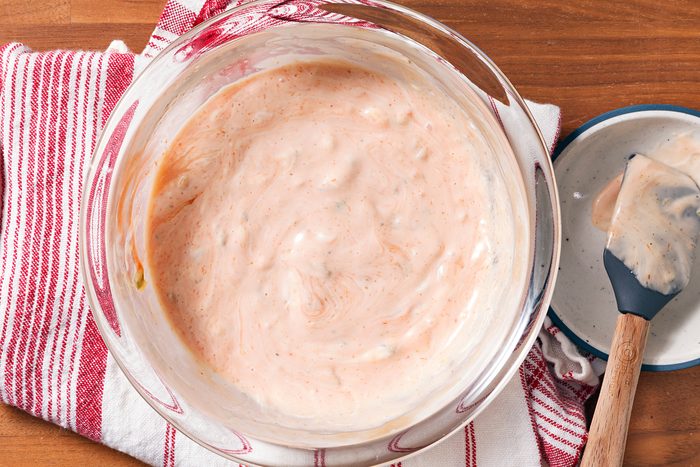 A bowl of dressing in a glass bowl on a red and white striped towel. A spatula with dressing rests on the side of the bowl. The creamy dressing has a light pink hue with visible herbs.