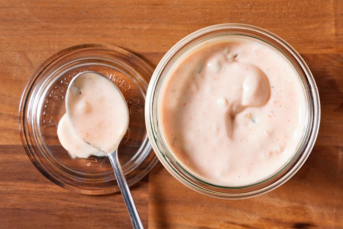 A top-down view of a glass jar filled with creamy Thousand Island dressing on a wooden surface. A spoon rests on the jar's lid beside it, with a dollop of dressing on the spoon.