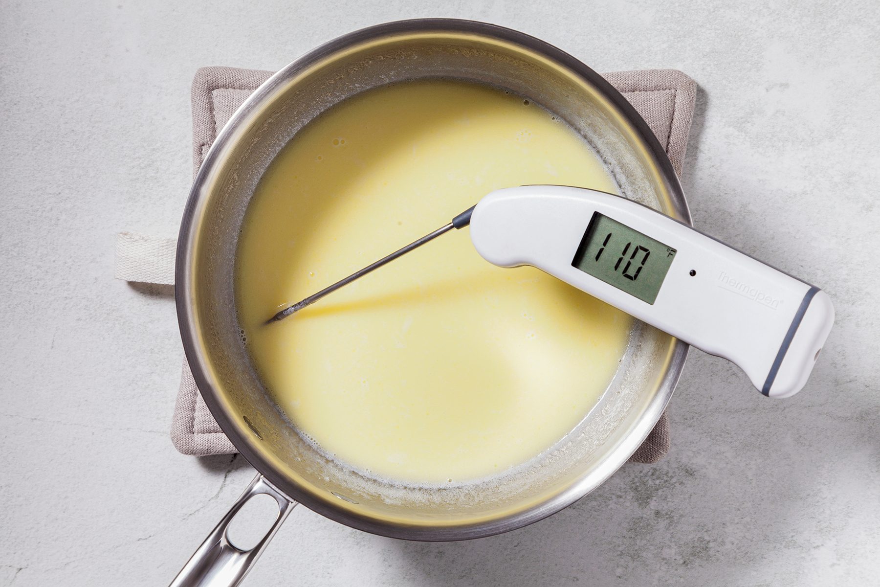 A stainless steel pot containing a creamy mixture is placed on a beige potholder. A digital thermometer inserted into the mixture reads 110 degrees. The surface surrounding the pot is light gray.
