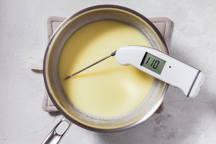 A stainless steel pot containing a creamy mixture is placed on a beige potholder. A digital thermometer inserted into the mixture reads 110 degrees. The surface surrounding the pot is light gray.