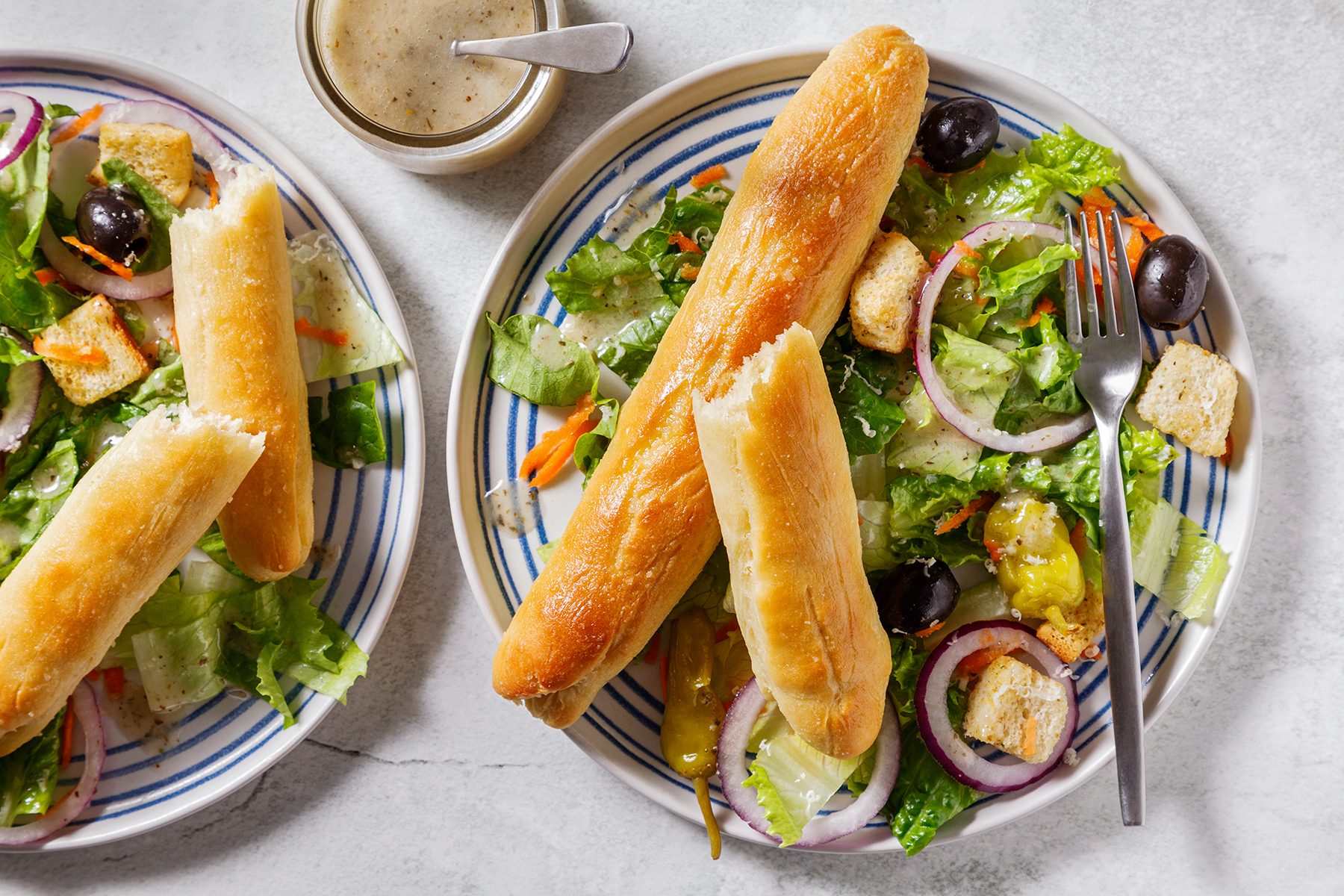 Two plates of salad with lettuce, sliced onions, olives, croutons, and breadsticks on striped plates. A small jar of dressing with a spoon is placed on the side.