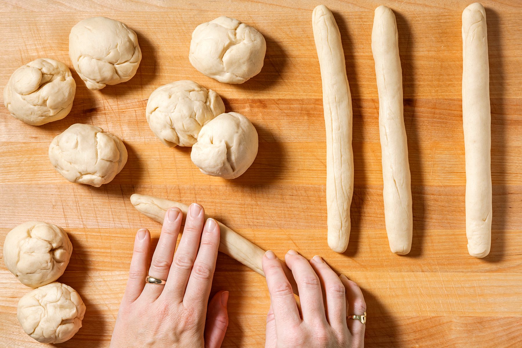 Hands shaping dough into long rolls on a wooden surface, with several dough balls nearby.
