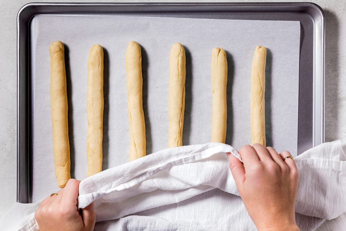 Hands are covering six breadsticks on a baking sheet lined with parchment paper using a white cloth.