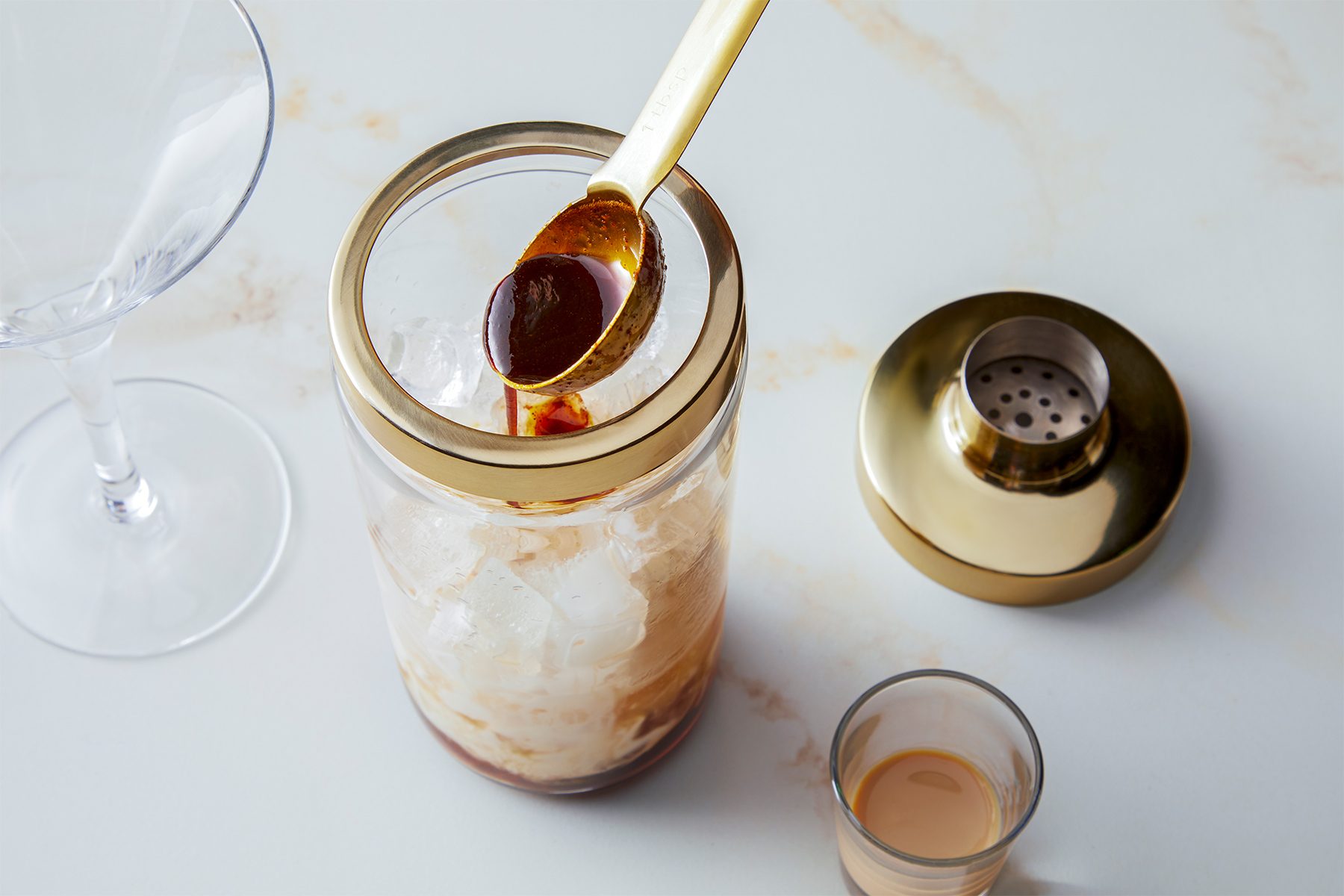 A gold cocktail shaker sits next to a glass filled with ice, with a golden spoon resting on its rim. A small shot glass with liquid is nearby, and the scene is set on a marble surface.