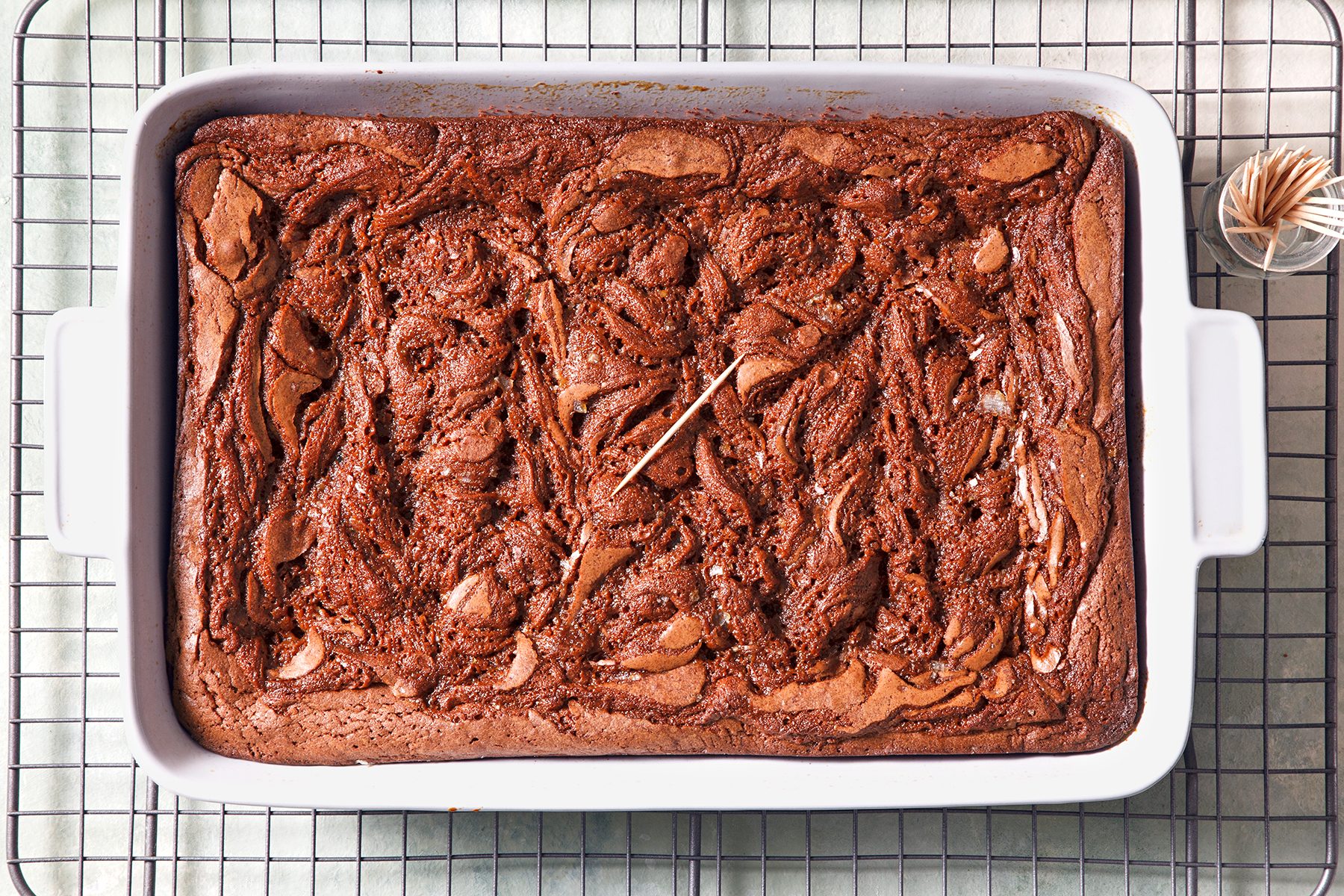 A rectangular baking dish filled with swirled chocolate brownies cooling on a wire rack. A toothpick is inserted in the center for doneness testing, and a small container of additional toothpicks is nearby.