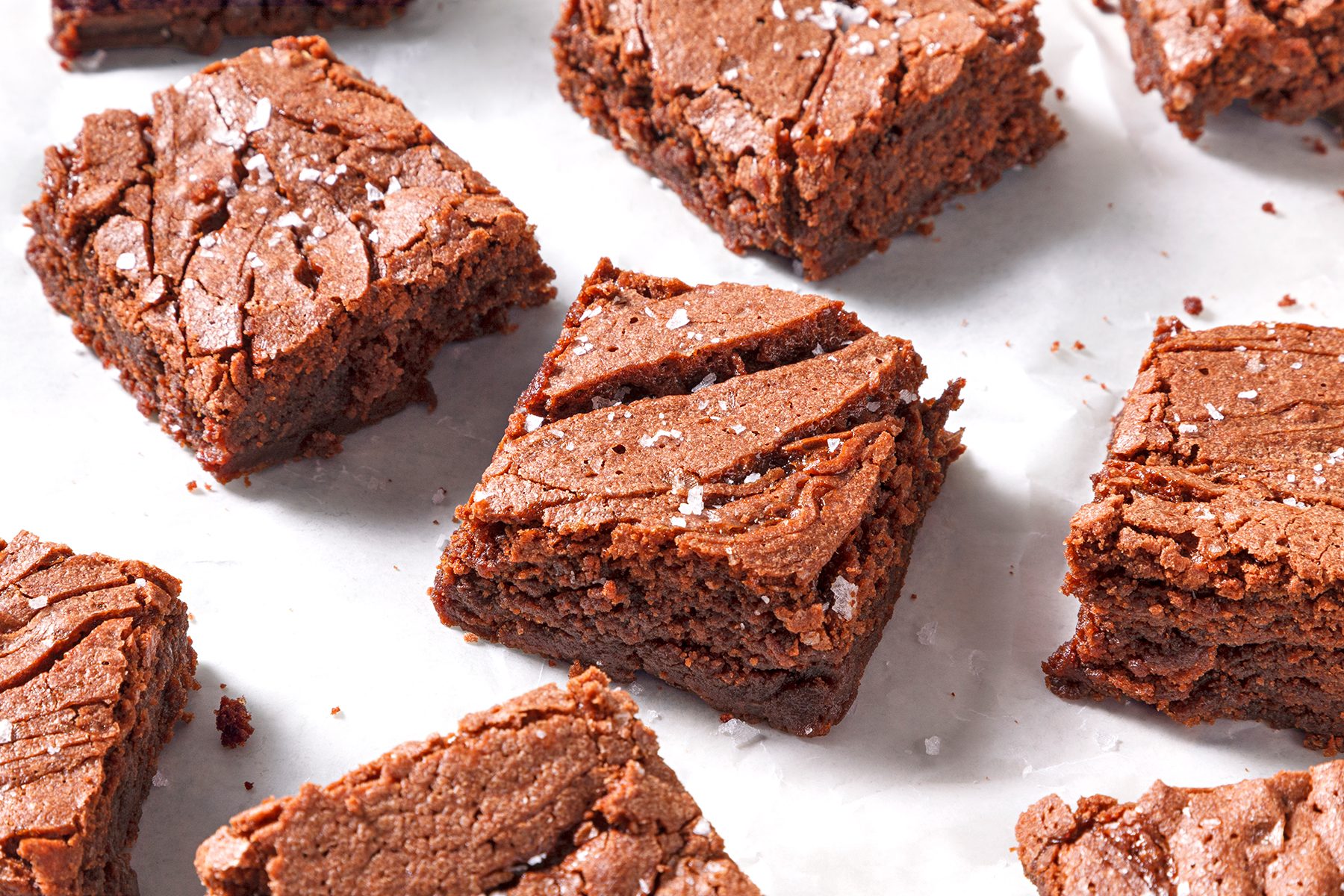 Squares of chocolate brownies sprinkled with sea salt are arranged on a white parchment paper. The brownies have a rich, fudgy texture with slightly cracked tops.