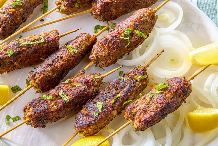 overhead shot of grilled kebab skewers, the skewers are arranged on a white plate with sliced onions and lemon wedges, the kebabs are cooked to a golden brown, they are topped with fresh herbs;