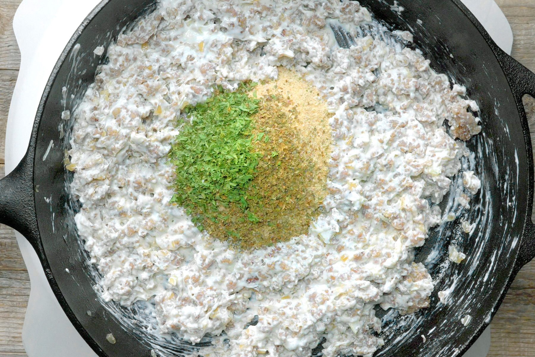 Skillet with creamy ground meat mixture topped with green herbs and breadcrumbs.