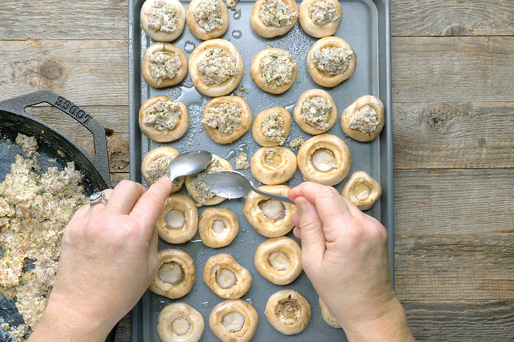 Hands fill mushroom caps with a stuffing mixture using two spoons on a baking tray. A pan with leftover stuffing mixture is visible on the side.