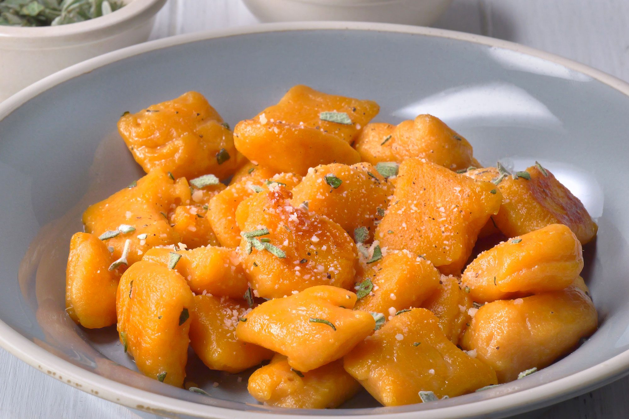 3/4th shot of a plate of pumpkin gnocchi; the gnocchi is cooked and golden brown and has been tossed in a sauce of butter, sage, and parmesan cheese; the gnocchi is in a light blue bowl, and the white wood background
